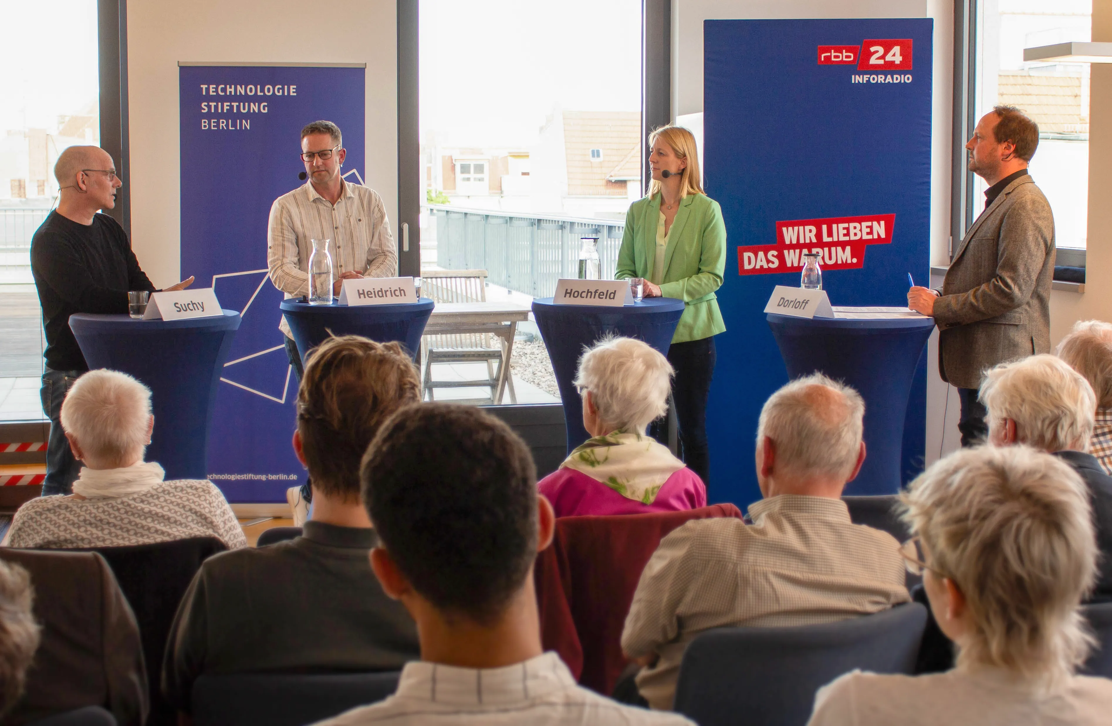 Vier Menschen mit Mikrofonen an Stehtischen, im Vordergrund sitzende Menschen, die der Diskussion zuhören. Im Hintergrund sind zwei Banner der Technologiestiftung Berlin und rbb24 Inforadio zu sehen.